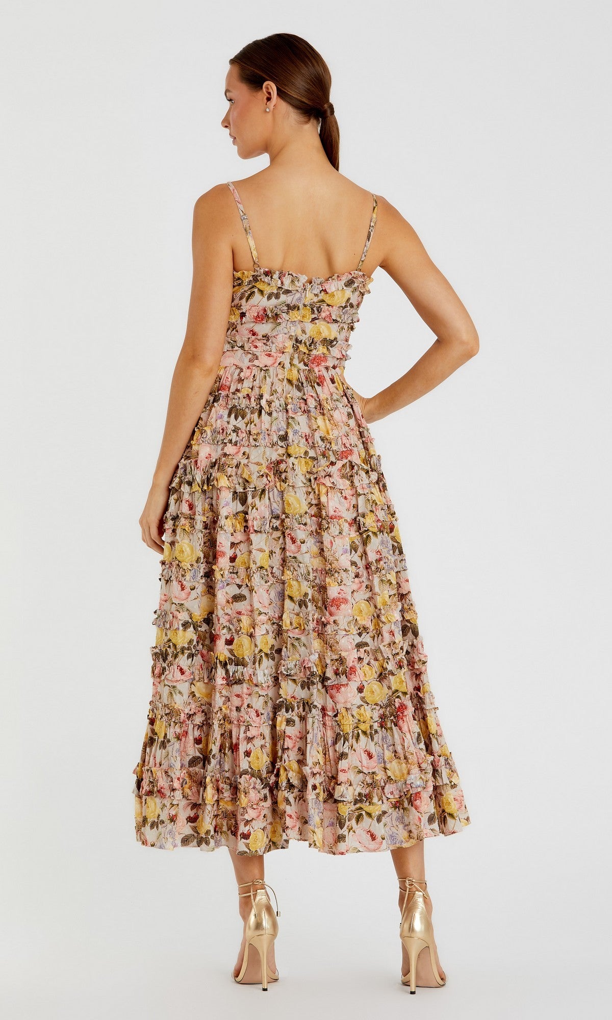 A woman faces away, wearing the Mac Duggal 8026 Midi Wedding Guest Dress with floral print and ruffles, paired with gold high heels, against a plain light background.